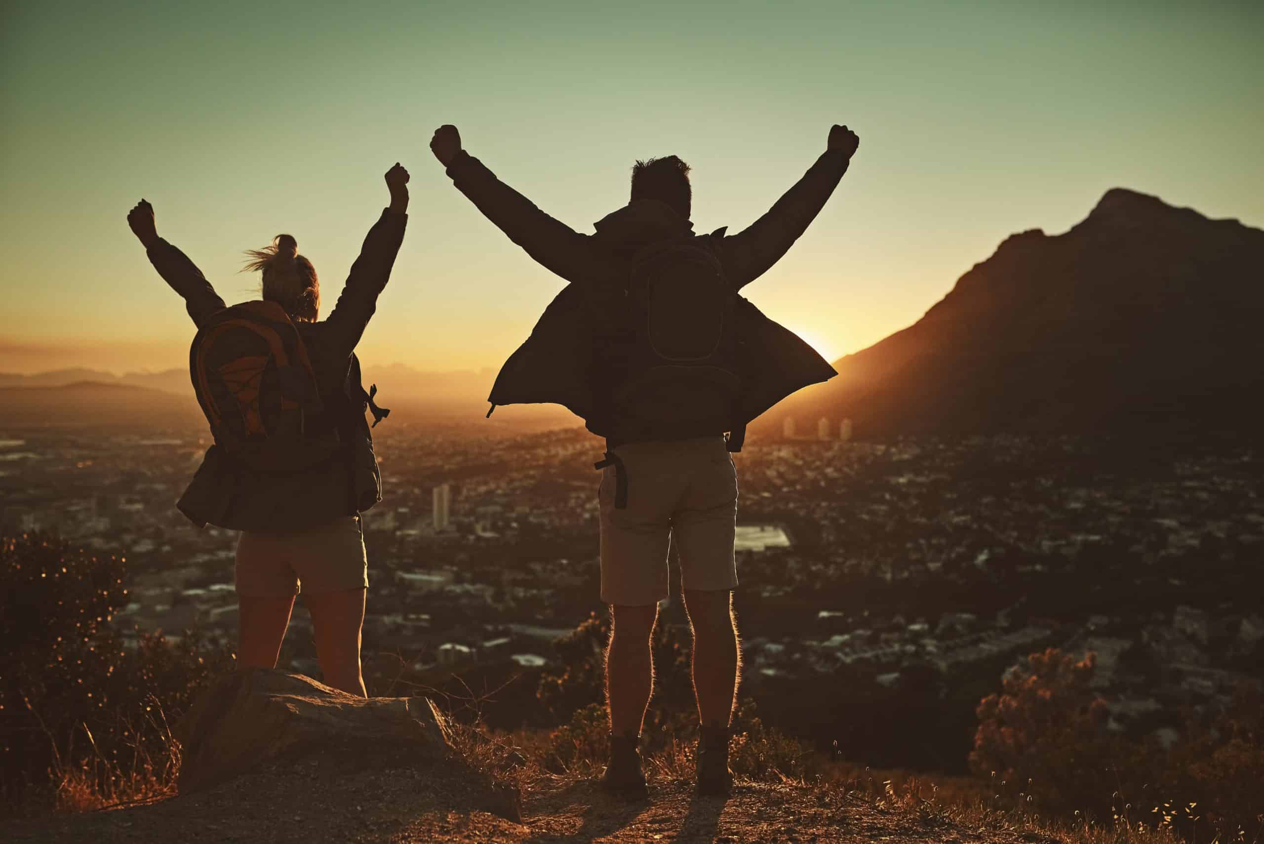 Verslavingszorg in Zuid-Afrika: Waar Moet Je Op Letten? Two hikers celebrating at sunrise on a mountain overlooking a cityscape in South Africa.
