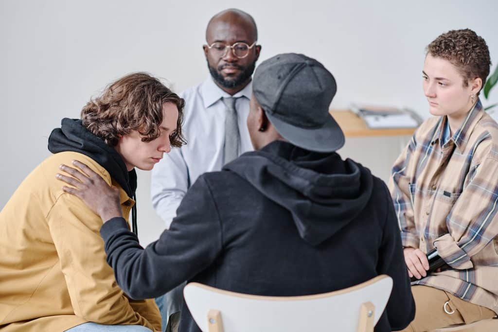 Groepstherapie waarbij een man troostend een hand op de schouder legt van een jongere cliënt, passend bij dubbele-diagnose behandeling.